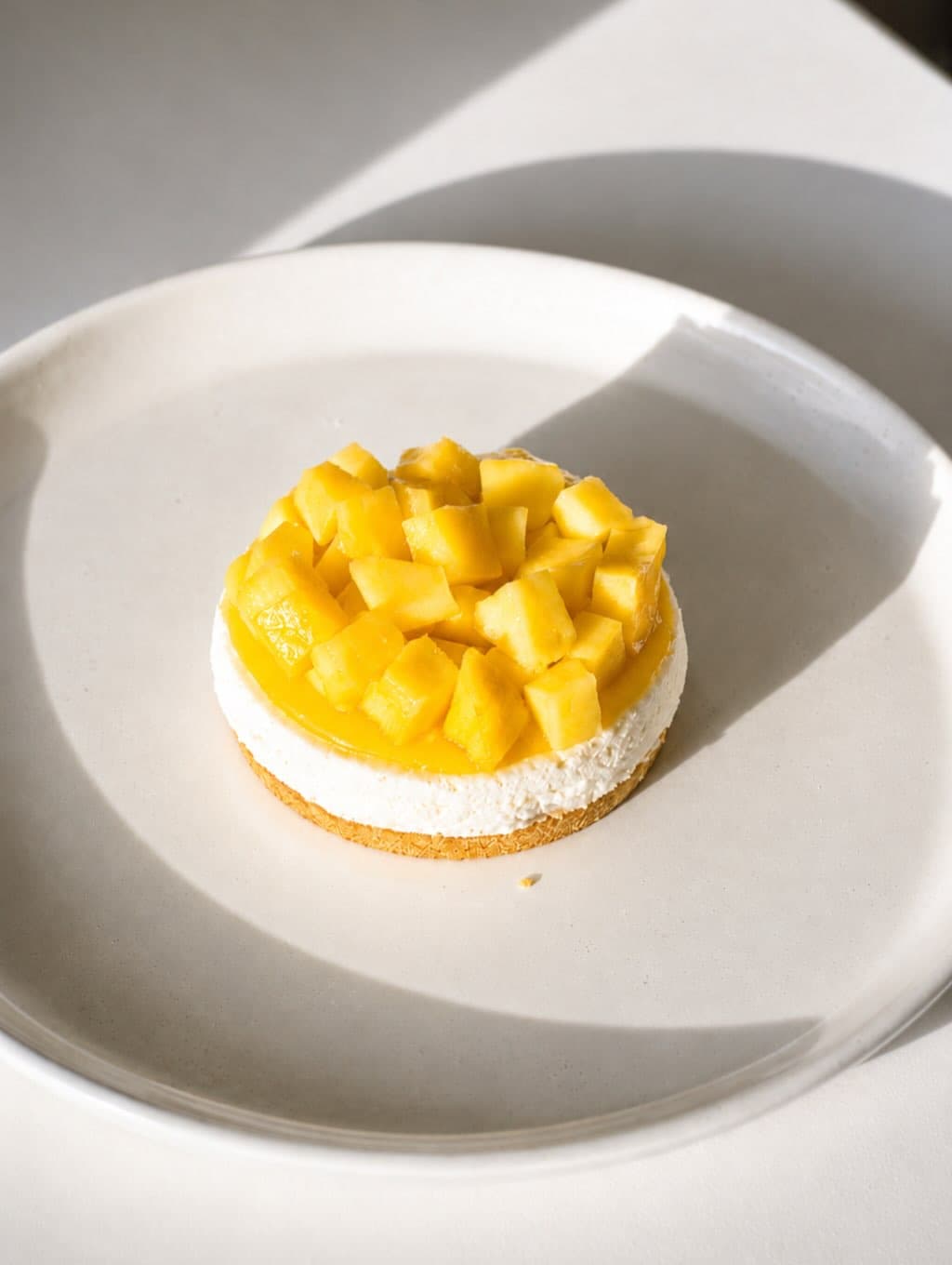A round mango-topped cheesecake on a white plate in hard daylight