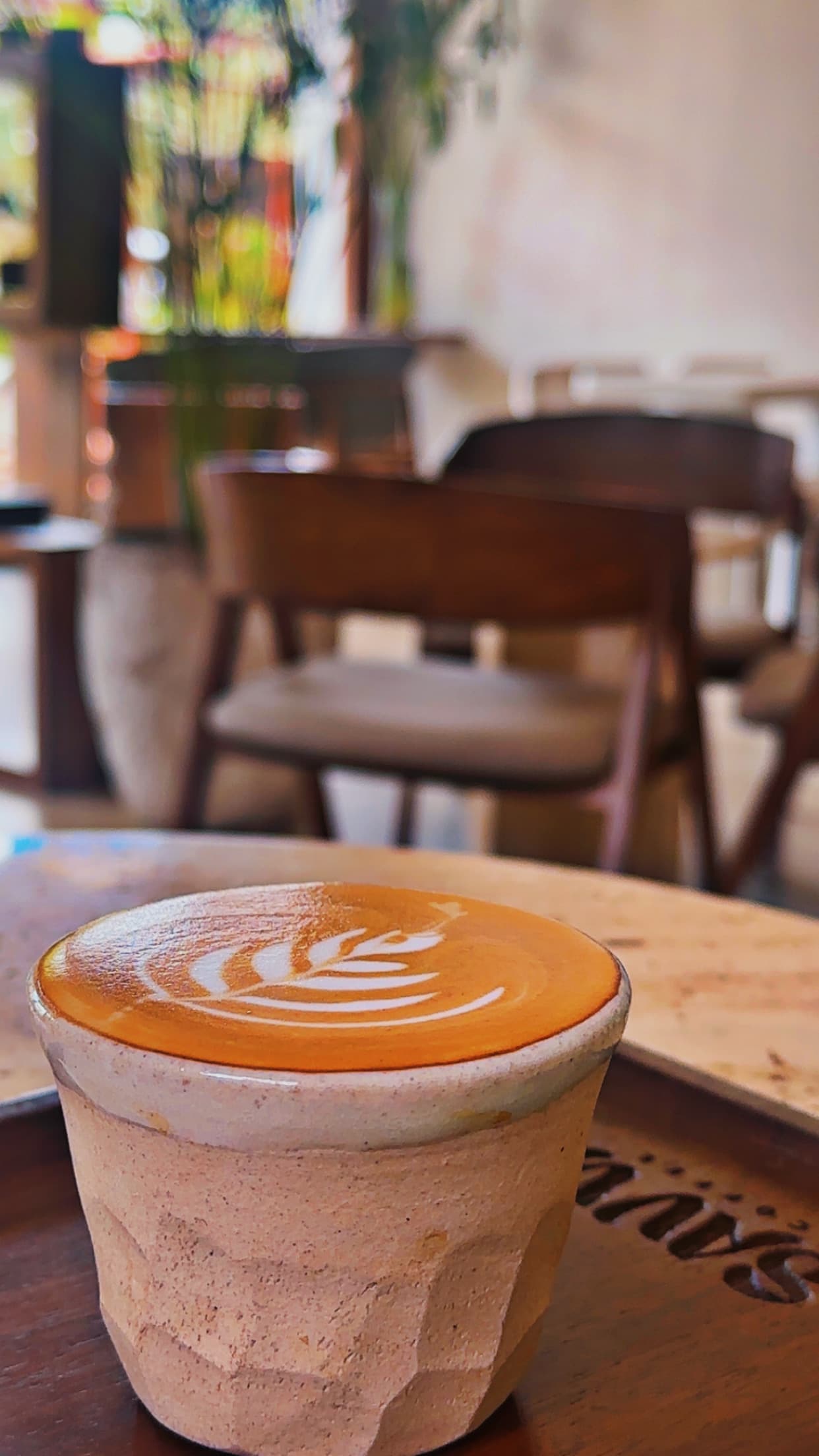 A latte with leaf-pattern art on a wooden table, SAVVA's wooden chairs and lounge softly out of focus behind